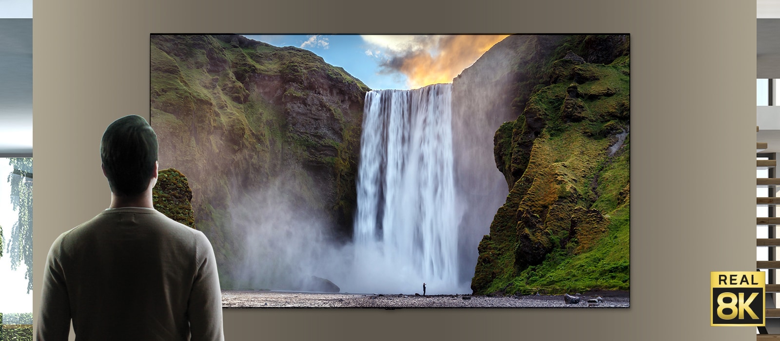 一个男人站在一个从悬崖流落的大瀑布前的壮观景色。场景缩小以显示该瀑布其实是安装在墙上的电视上的图像。