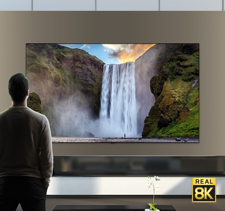 一个男人站在一个从悬崖流落的大瀑布前的壮观景色。场景缩小以显示该瀑布其实是安装在墙上的电视上的图像。