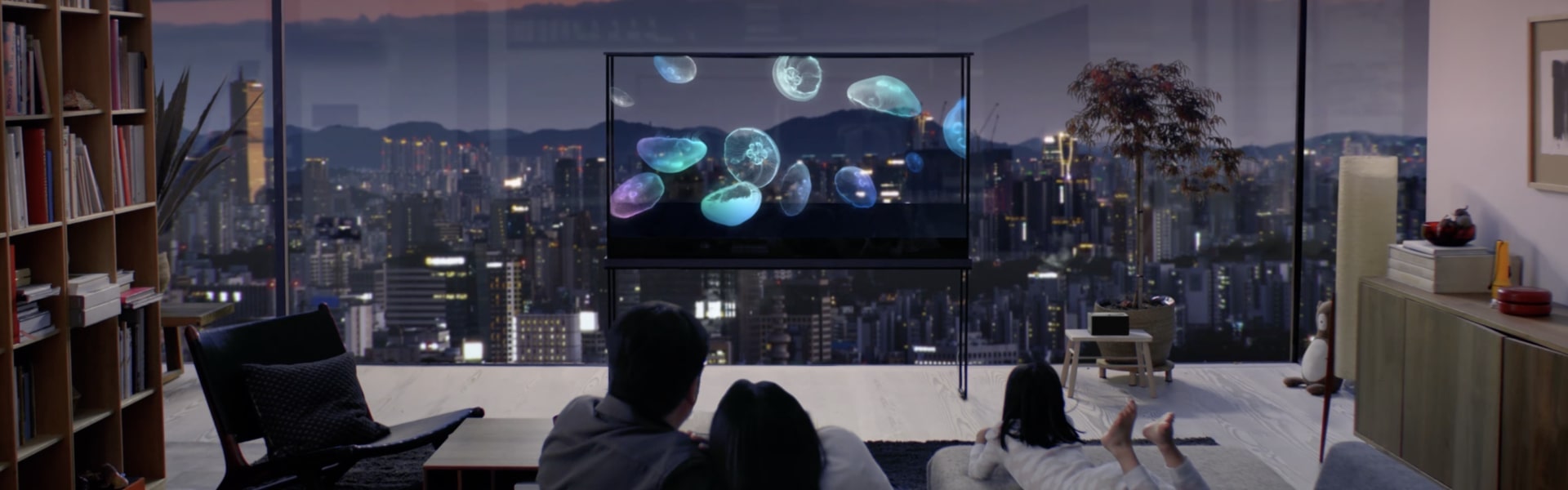 一家人正在享受一台 LG Oled T，上面漂浮着一只水母，而城市景观则透过透明屏幕映入眼帘。