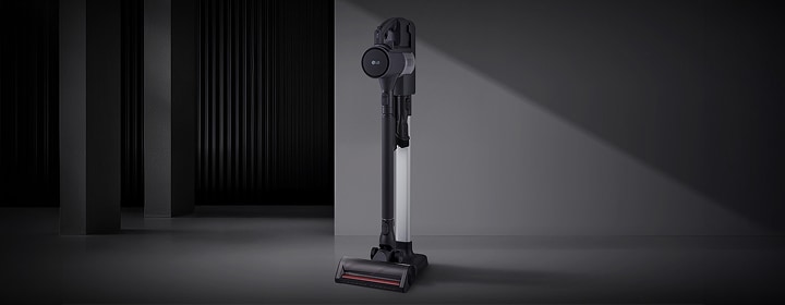 LG cordless stick vacuum cleaner in matte black and silver, upright on a dark gray studio floor with shadowed wall.