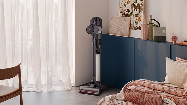LG cordless stick vacuum in black and silver upright beside blue cabinet, sheer white curtains, and a bed with pink quilt.