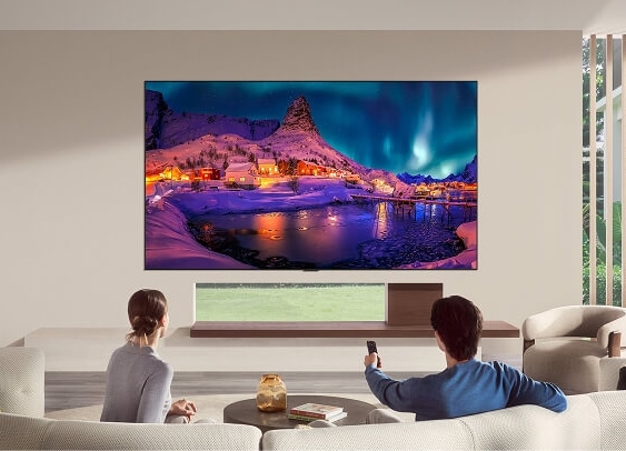 LG OLED evo AI G6 is flush-fit wall-mounted in a modern living room, displaying a vivid aurora over a snowy coastal village while a man and woman watch from the sofa with a remote in hand.