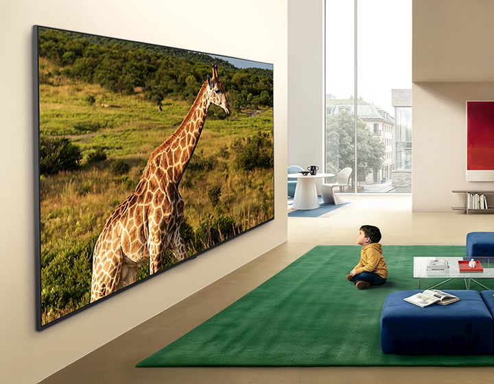 Le téléviseur LG QNED evo AI QNED85 Mini LED Ultra Big est fixé au mur dans un espace de vie lumineux, affichant une girafe dans un paysage naturel avec des détails éclatants qui mettent l’accent sur une échelle immersive et une forte présence pour un enfant assis à proximité.