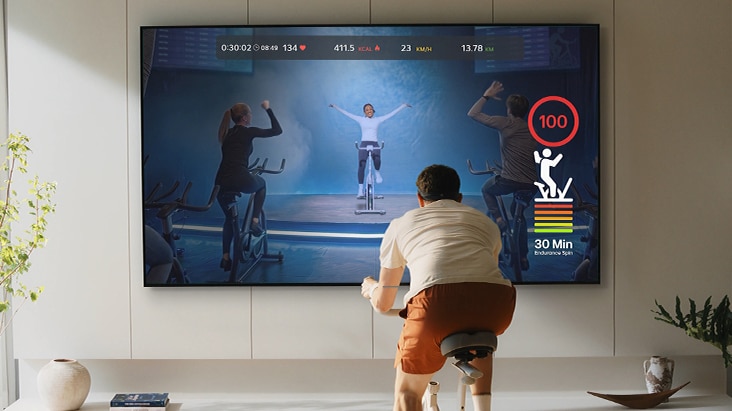 Le téléviseur LG AI, doté du service de conditionnement physique Fitness Machine, montre un utilisateur s’entraînant sur un vélo stationnaire tandis qu’un écran de télévision affiche une séance d’entraînement en direct avec des mesures de conditionnement physique en temps réel, des visuels d’entraînement et un environnement de conditionnement physique immersif à domicile.