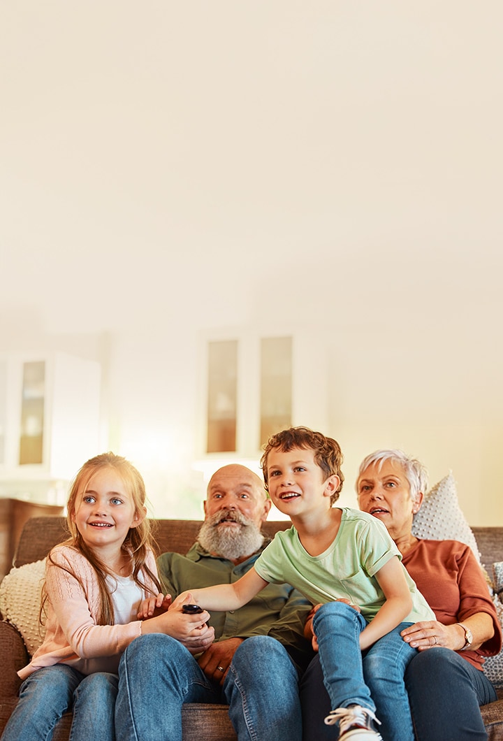 Eine Familie mit Kindern und ihren Großeltern sitzt zusammen auf einem Sofa in einem hellen Wohnzimmer und hält eine Fernbedienung, während sie fernsieht.