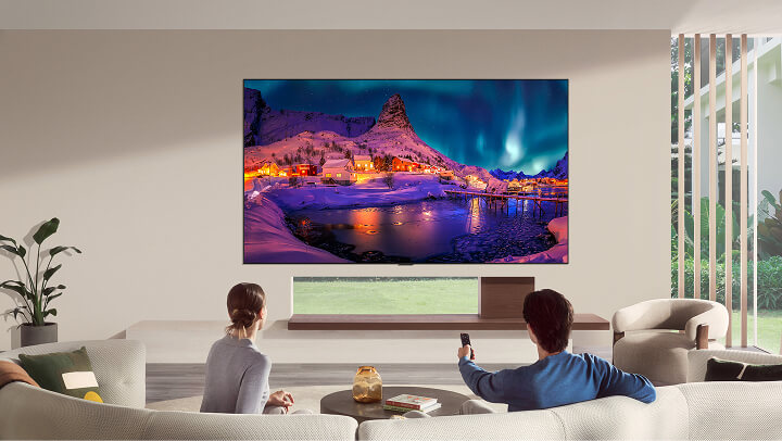 Der LG OLED evo AI G6 ist in einem modernen Wohnzimmer bündig an der Wand montiert und zeigt eine lebhafte Aurora über einem verschneiten Küstendorf, während ein Mann und eine Frau vom Sofa aus mit einer Fernbedienung in der Hand zusehen.