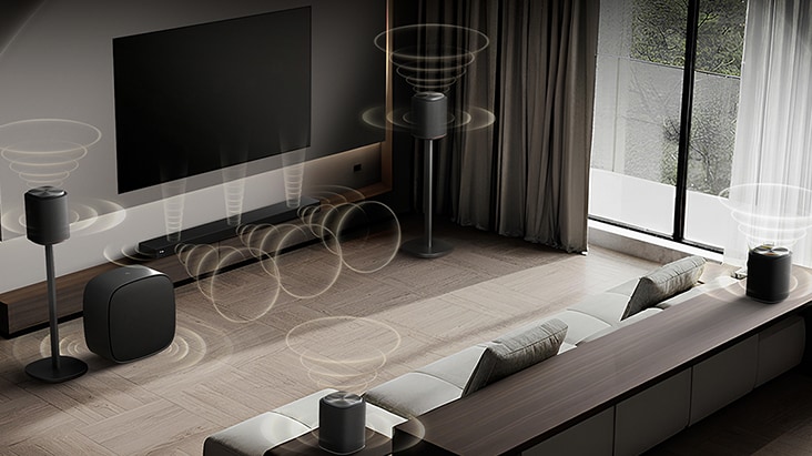 LG TV se Sound Suite je nástěnná v moderním obývacím pokoji, s několika reproduktory uspořádanými kolem pohovky, přičemž vizualizované zvukové vlny vyzařují ven, což ilustruje flexibilní zvukové nastavení přizpůsobené prostoru.