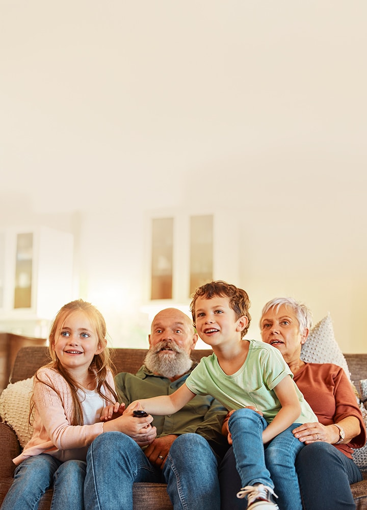 Eine Familie mit Kindern und ihren Großeltern sitzt zusammen auf einem Sofa in einem hellen Wohnzimmer und hält eine Fernbedienung, während sie fernsieht.
