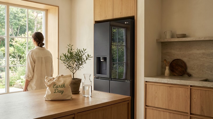LG refrigerator with sleek glass panel in a natural wood kitchen, beside a large window with greenery and natural light.	