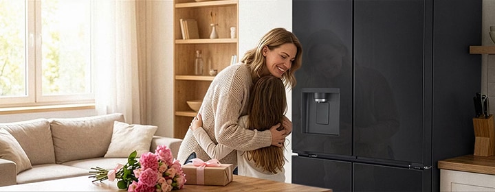 A mother and a child embracing next to a stainless-steel LG refrigerator, beside a bouquet of flowers and a gift on a table.	