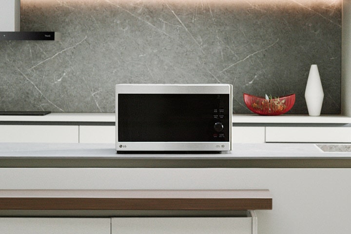 A silver countertop microwave sits in a modern kitchen with a gray stone backsplash, a sink, and a built-in oven.