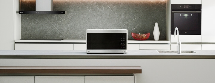 A silver countertop microwave sits in a modern kitchen with a gray stone backsplash, a sink, and a built-in oven.