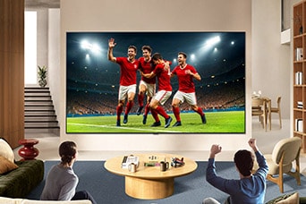 LG TV for sports fans is wall-mounted in a bright living room, showing a soccer celebration with vivid colors and refined picture quality across a wide screen, as a family seated on sofas cheers together.