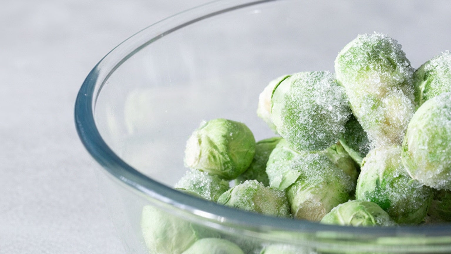 Una ciotola di vetro piena di cavoli di Bruxelles surgelati leggermente coperti di gelo, posizionati su una superficie grigio chiaro.
