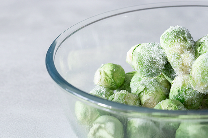 Una ciotola di vetro piena di cavoli di Bruxelles surgelati leggermente coperti di gelo, posizionati su una superficie grigio chiaro.