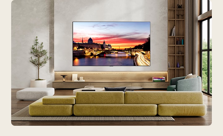Телевизор LG OLED evo AI C6 закреплен на стене в стильной гостиной, демонстрируя ультратонкую конструкцию с узкими рамками, и на экране – изображение городского пейзажа у реки на закате. 