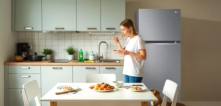 Réfrigérateur-congélateur LG à 2 portes rose et noir dans une cuisine moderne. Une femme prend un petit-déjeuner à table avec de la nourriture.