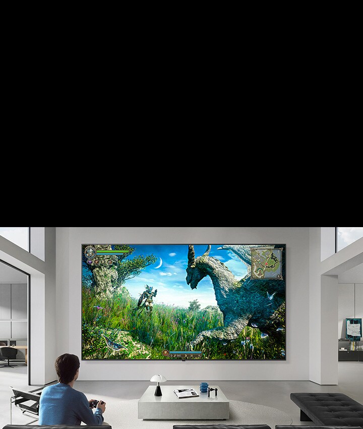 Le LG TV apparaît monté sur un mur dans un salon aux tons noir et blanc, affichant une scène de jeu de dinosaures fantastique sur un grand écran tandis qu'une personne est assise sur un canapé tenant une manette.