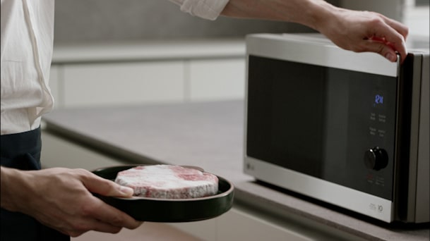 Una persona abre la puerta de un microondas LG plateado con una mano mientras sostiene un plato con carne cruda junto a la encimera.