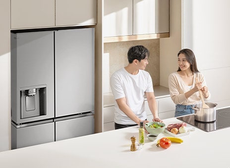 A large stainless LG refrigerator freezer with spacious storage in a stylish kitchen. A man and a woman cook in front of a table.