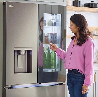 A woman in a pink shirt taps the glass panel of an LG InstaView fridge. Through the transparent panel, water bottles and groceries inside the fridge are visible.