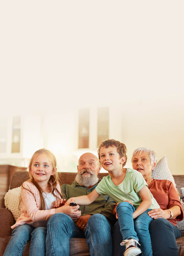 En familj med barn och deras mor- och farföräldrar sitter tillsammans i en soffa i ett ljust vardagsrum och håller en fjärrkontroll medan de tittar på TV.