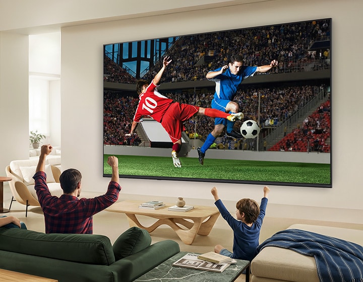 LG QNED AI QNED70 Mini LED ultrastor TV väggmonterad i ett ljust vardagsrum visar ett intensivt fotbollsögonblick med intensiva färger och förfinad bildkvalitet över en bred skärm, medan en familj som sitter i soffor hejar tillsammans.