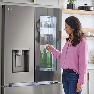 A woman in a pink shirt taps the glass panel of an LG InstaView fridge. Through the transparent panel, water bottles and groceries inside the fridge are visible.