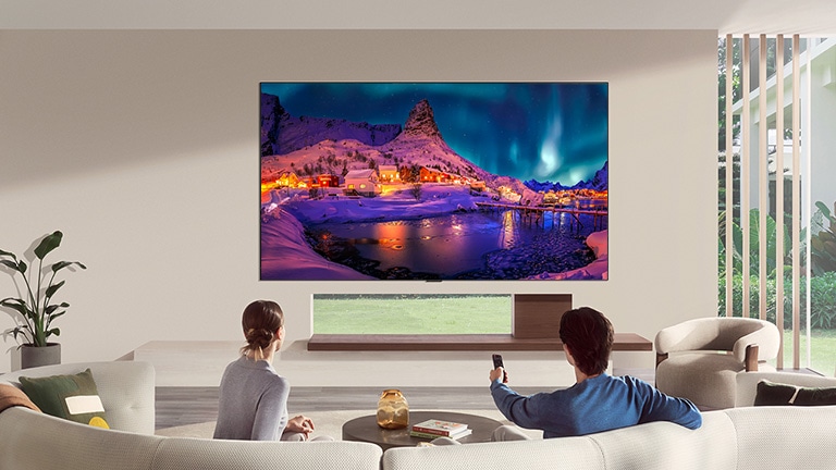 LG OLED evo AI G6 is flush-fit wall-mounted in a modern living room, displaying a vivid aurora over a snowy coastal village while a man and woman watch from the sofa with a remote in hand.