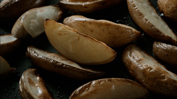 A cooking close-up shows golden potato wedges with browned skins and glossy surfaces on a dark pan.