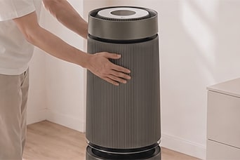 Hình ảnh một người dùng cả hai tay chạm hoặc điều chỉnh lớp vỏ ngoài có bề mặt họa tiết của máy lọc không khí hình trụ cao, màu xám đậm.