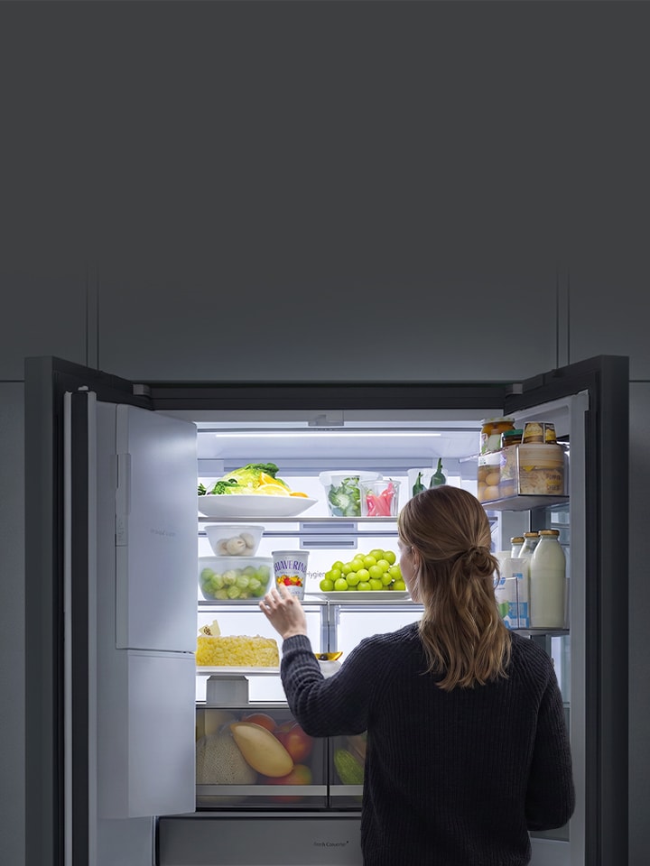 Ảnh chụp từ phía sau một người đang với tay lấy đồ trong tủ lạnh LG, với hệ thống đèn Full Back chiếu sáng rõ ràng các thực phẩm bên trong.