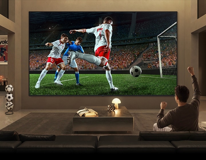 LG Micro RGB evo TV với màn hình 100 inch cung cấp chế độ xem thể thao đắm chìm, hiển thị một trận bóng đá quy mô lớn với các cầu thủ chân thực và chi tiết sân vận động lấp đầy màn hình trong khi người xem nâng một tay lên để ăn mừng.