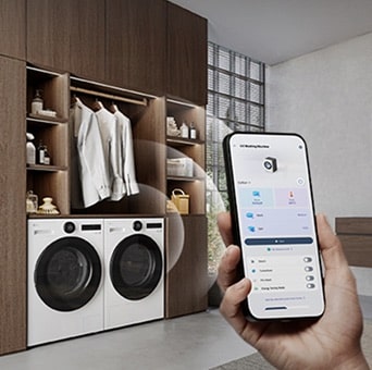 Person holding a smartphone with the ThinQ app open, controlling front load washers in a laundry room.