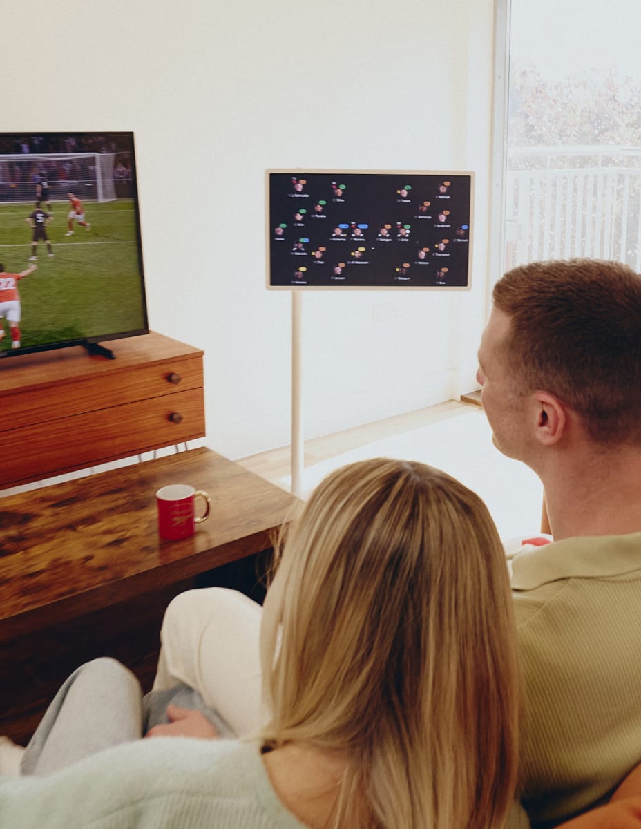 Die Rückansicht eines Mannes und einer Frau, die nebeneinander auf einem Sofa sitzen und ein Fußballspiel im TV schauen, während sie eine Spielanalyse auf einem StanbyME Smart Screen verfolgen.