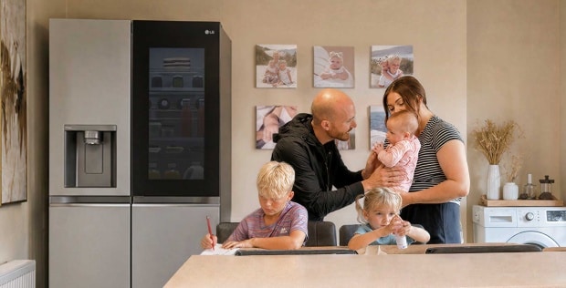 Eine Familie versammelt sich in einer Küche neben einem Edelstahl-LG-Kühlschrank – ein Junge schreibt am Esstisch, ein junges Mädchen sitzt neben ihm mit einer kleinen Flasche, und Eltern stehen zusammen und halten ein Baby.