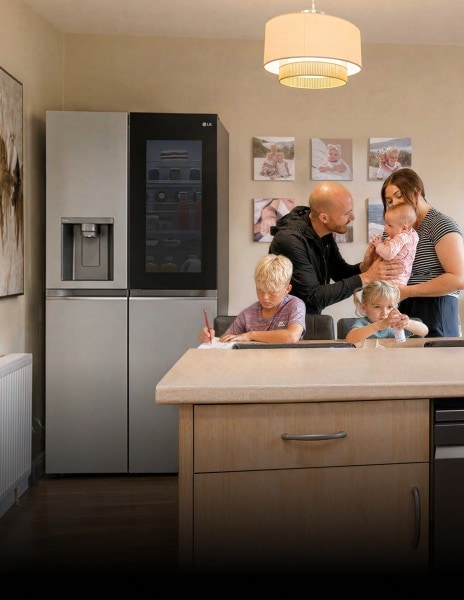Eine Familie versammelt sich in einer Küche neben einem Edelstahl-LG-Kühlschrank ein Junge schreibt am Esstisch, ein junges Mädchen sitzt neben ihm mit einer kleinen Flasche, und Eltern stehen zusammen und halten ein Baby.