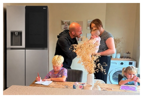Eine Familie versammelt sich in der Küche neben einem Edelstahl-Kühlschrank – ein Junge zeichnet am Tisch, ein Mädchen malt an der Arbeitsfläche, und die Eltern stehen zusammen und halten ein Baby.