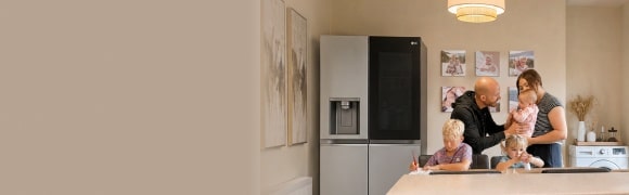 Eine Familie versammelt sich in einer Küche neben einem Edelstahl-LG-Kühlschrank – ein Junge schreibt am Esstisch, ein junges Mädchen sitzt neben ihm mit einer kleinen Flasche, und Eltern stehen zusammen und halten ein Baby.
