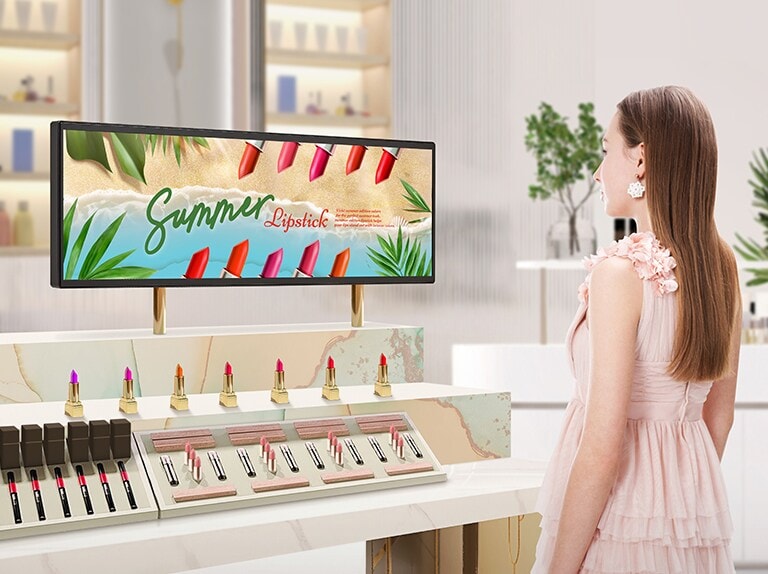 In einem hellen Kosmetikgeschäft ist ein BH7N in der Lippenstiftabteilung installiert. Eine Frau sieht sich eine anschauliche Werbung an, die auf dem Breitbildschirm des BH7N angezeigt wird.