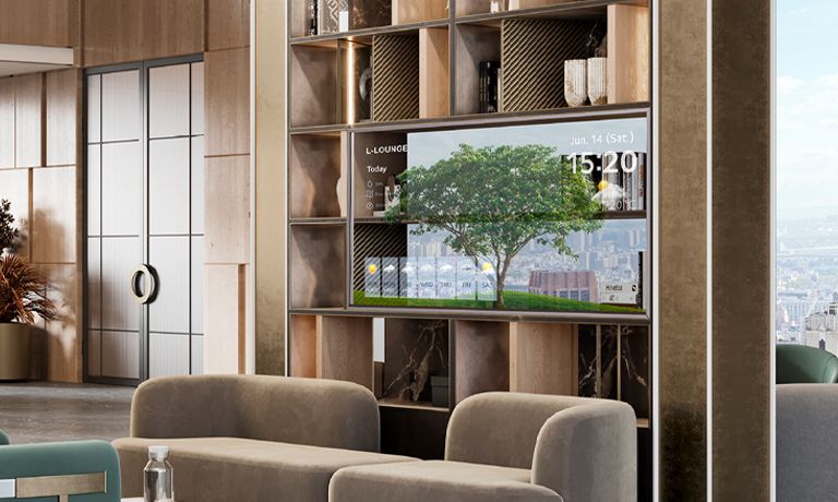 In der großen Lounge befindet sich eine offene Bildschirmeinheit mit einem transparenten OLED-Bildschirm in der Mitte. Auf dem Bildschirm sind Bilder von Bäumen sowie die aktuelle Uhrzeit und Wetterinformationen zu sehen. Die durch den transparenten Bildschirm sichtbare Landschaft lässt den Raum größer wirken.