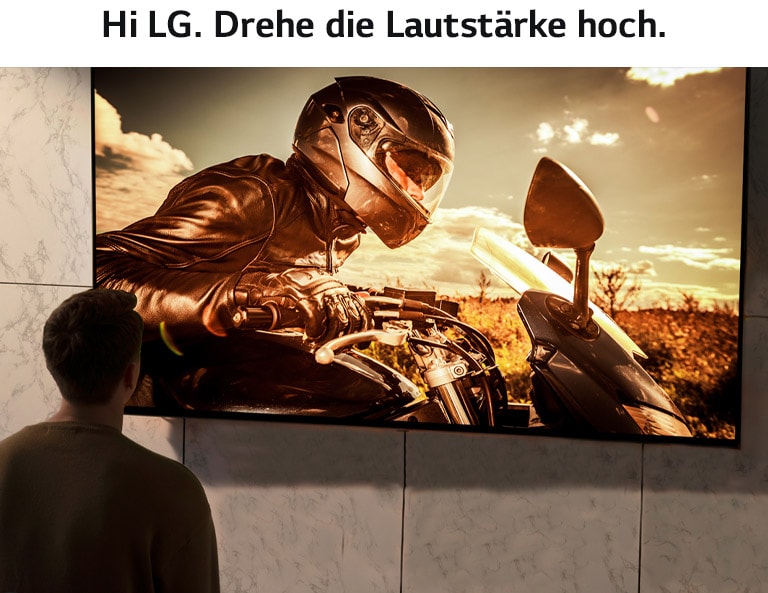 Ein Mann ist von hinten zu sehen und verfolgt das Geschehen auf einem wandmontierten Fernseher. Auf dem Bildschirm wird eine Filmszene mit Motorrädern wiedergegeben.