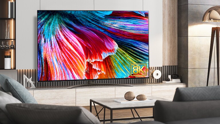 Der QNED99 steht an einer Wand in einem hell eingerichteten Raum. Vor dem Fernseher stehen Sofas und ein Couchtisch. Der Bildschirm zeigt eine Nahaufnahme von bunten Blütenblättern und das 8K-Logo.