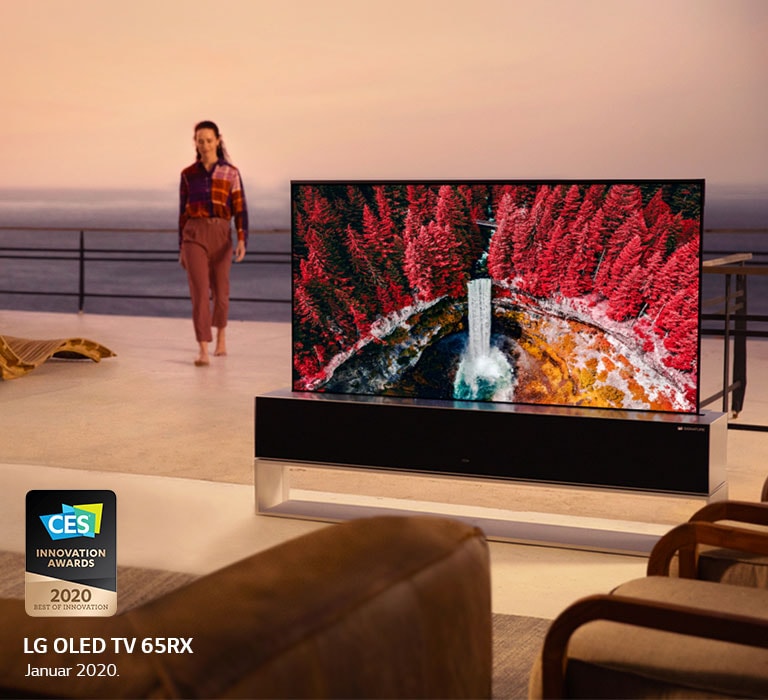 Wohnzimmer mit einem aufrollbaren Fernseher, der einen Wasserfall zwischen Ahornbäumen zeigt, während eine Frau von der Terrasse aus ins Haus geht