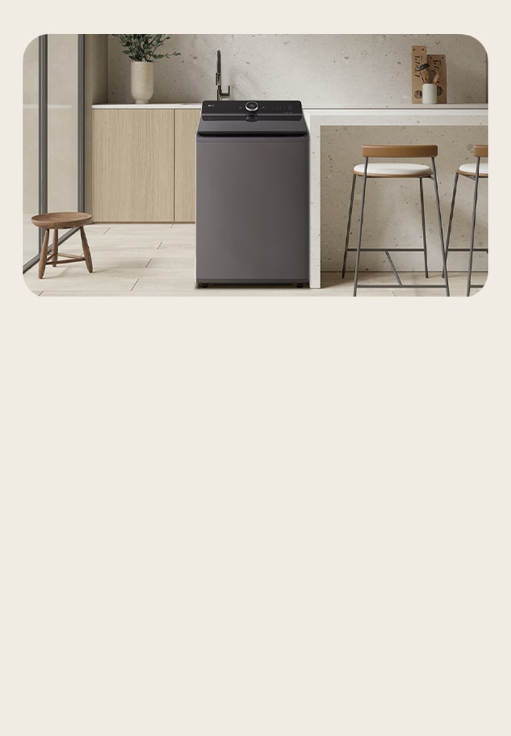 The top loading washing machine is located in the middle of a modern light brown interior. There is a large window on the left and a high stool on the right	