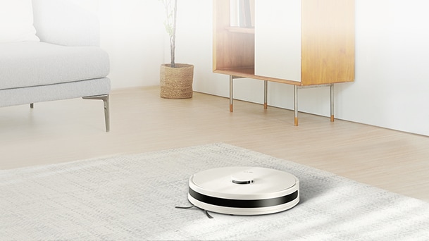 LG robot vacuum cleaner in white on a light rug in a bright living room with sofa and wood cabinet.