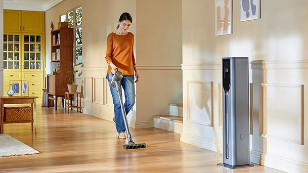 Person is using LG CordZero stick vacuum on a wood floor beside a tall gray All-in-One Tower in a warm home.