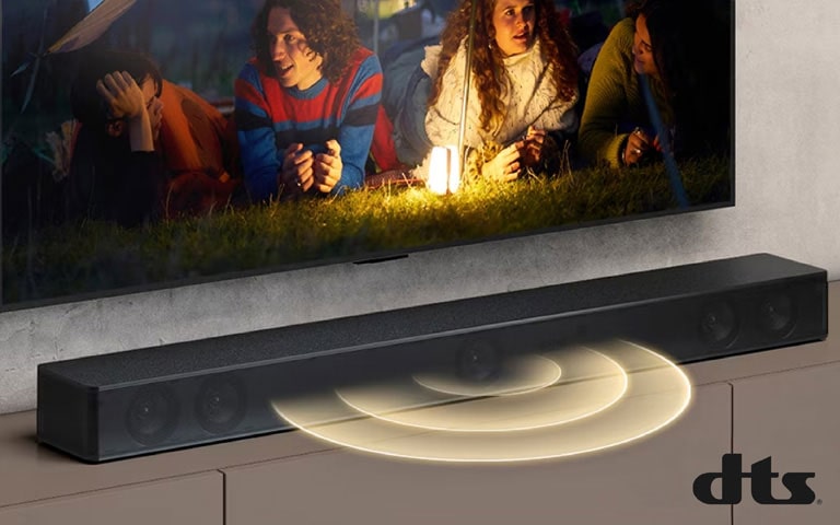 LG TV is on the wall, on the screen it shows 2 couples lying on the grass. In front of them, there is a lamp. LG Sound bar is below LG TV. Sound graphic is coming out from the front of the sound bar. DTS Virtual:X logo is shown on the bottom right corner of image.