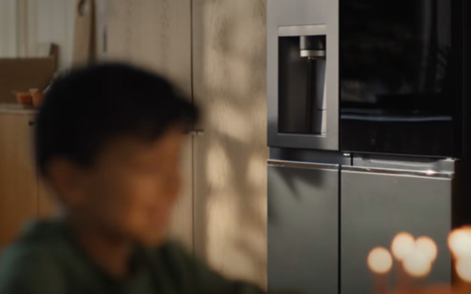 An out-of-focus child is in the centre of a kitchen. Behind him is a fridge and wooden storage drawers.