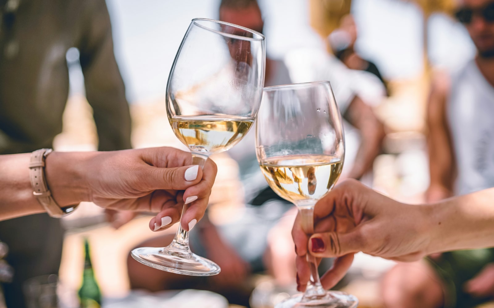 Two people clinking glasses of white wine in a celebratory toast outdoors, with a blurred background of friends and a sunny setting.